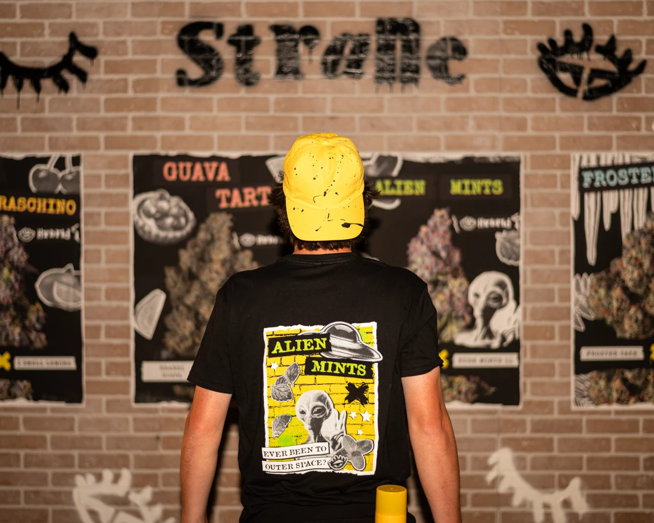 Black Strane Alien Mints graphic T-shirt and yellow cap, photographed against a poster-covered brick wall